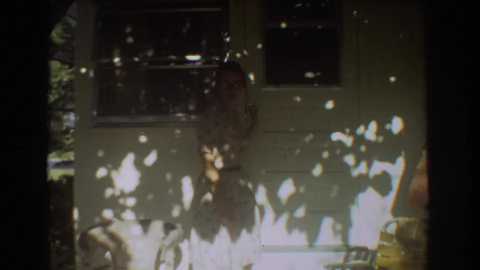 The frame shows a woman in a floral dress near the house exterior, with tree shadows and soft 8mm home-movie focus.