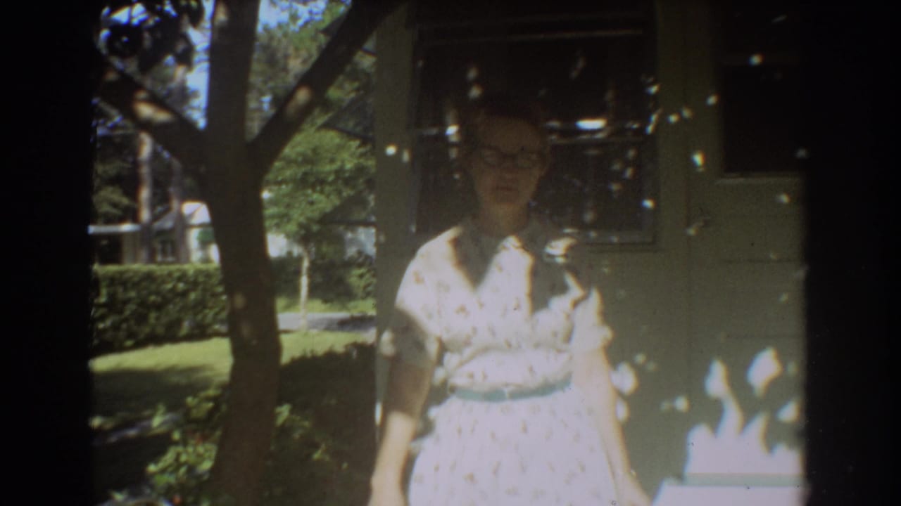 In 1967 Fort Lauderdale home-movie footage, a woman with glasses stands on a sun-dappled porch near a residential wall.