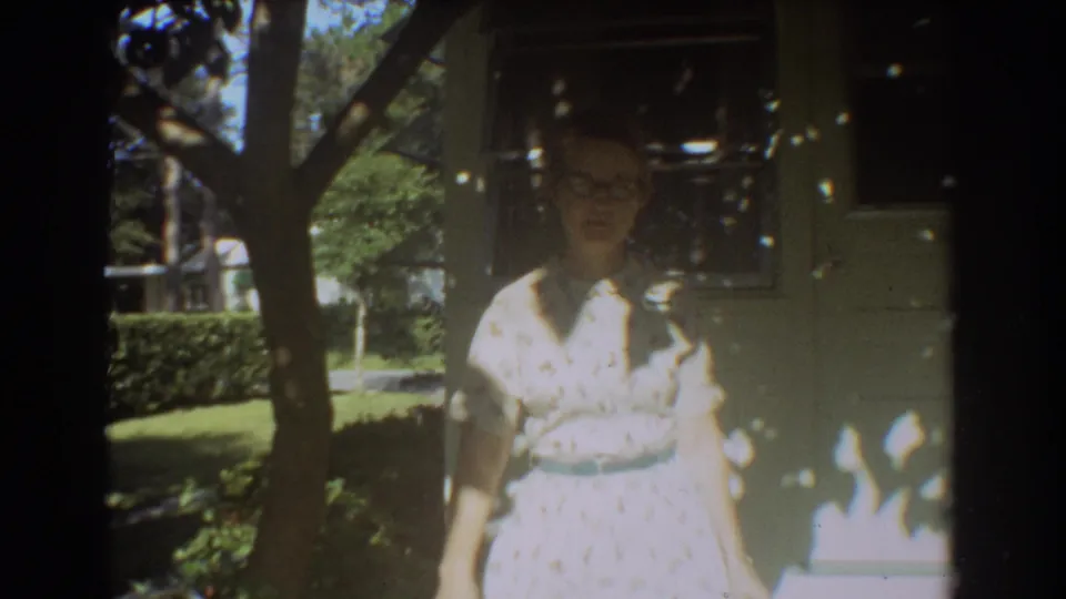 In 1967 Fort Lauderdale home-movie footage, a woman with glasses stands on a sun-dappled porch near a residential wall.
