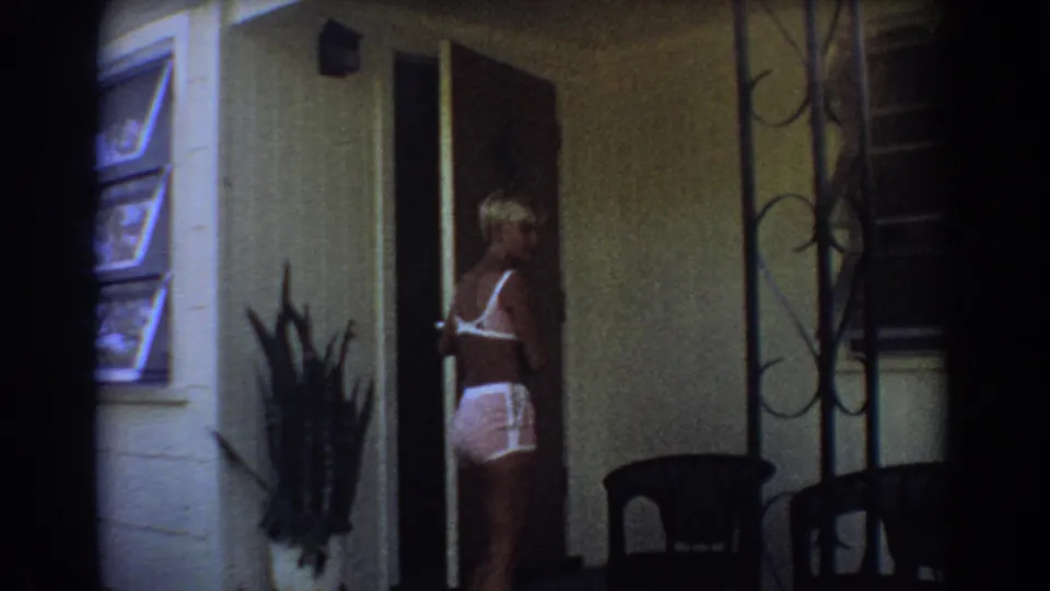 The blonde woman steps out through the doorway, with louvered windows and porch furniture visible around her.