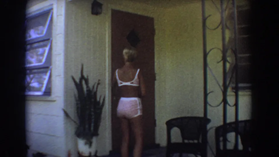 In 1967 Fort Lauderdale home-movie footage, a blonde woman in pink clothing faces a front door near a jalousie window and porch plants.