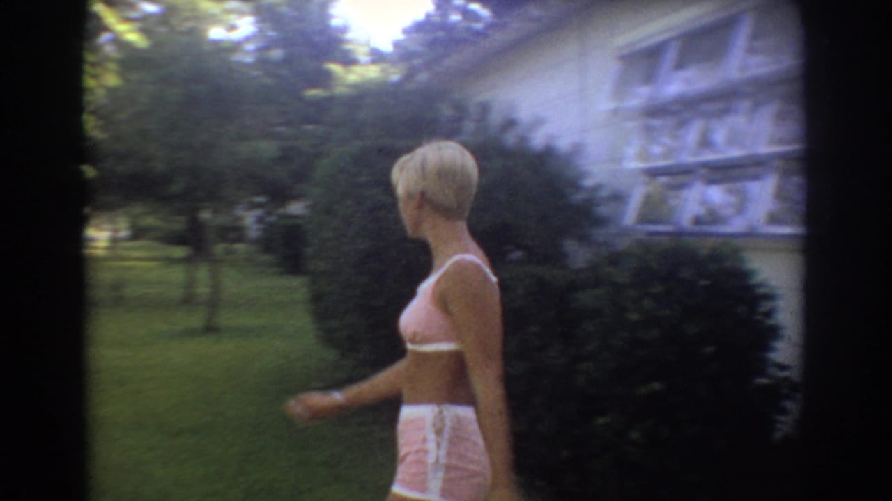 The woman walks through a sunlit residential yard in Fort Lauderdale, with shrubs, lawn, and house windows behind her.