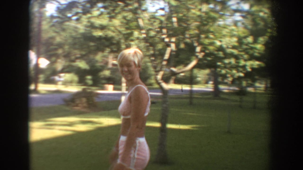 In the Fort Lauderdale yard, the blonde woman turns and smiles toward the home-movie camera with lawn, trees, and a street area behind her.
