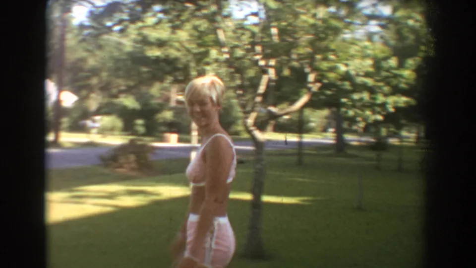 In the Fort Lauderdale yard, the blonde woman turns and smiles toward the home-movie camera with lawn, trees, and a street area behind her.