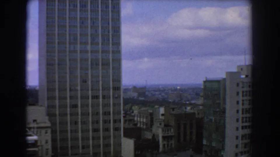 High-angle Melbourne city view from 1967 8mm footage, showing a tall tower, urban buildings, horizon, and clouded sky.