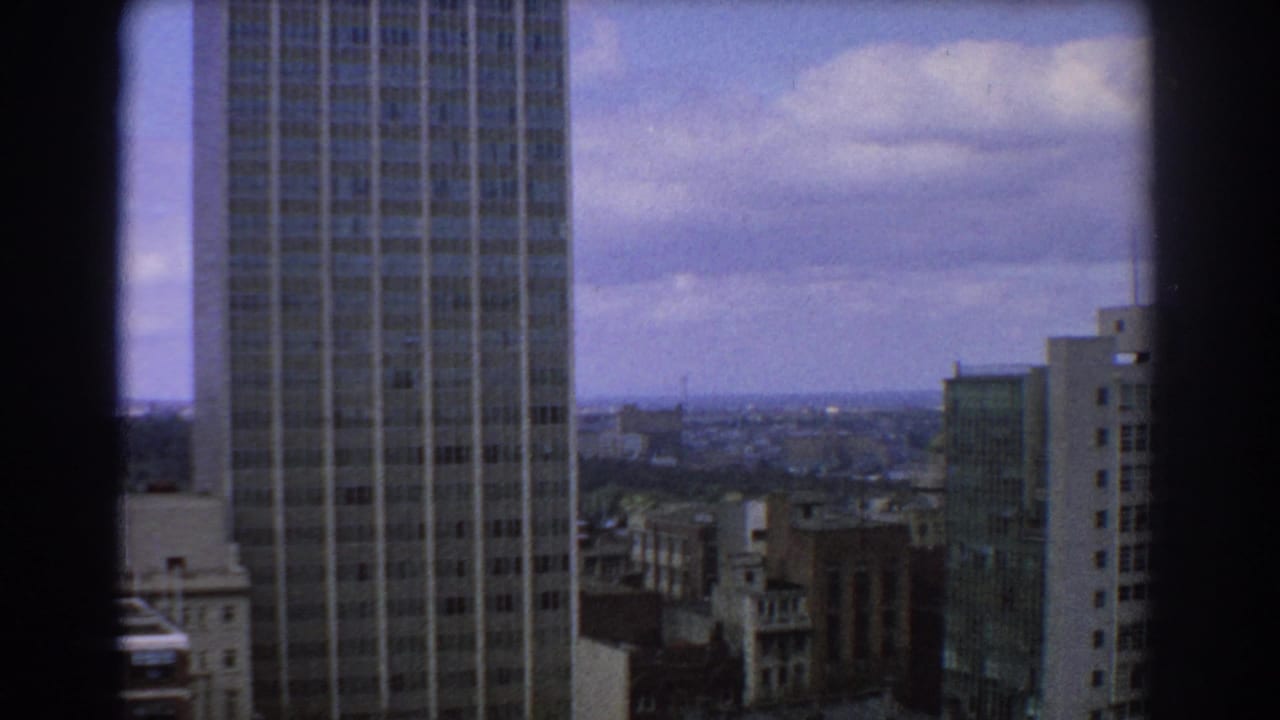 High-angle city view with a modern grid-faced tower and distant buildings under clouds.