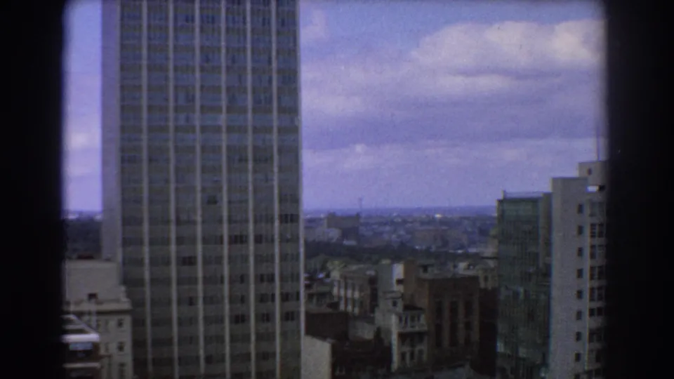 1967 Melbourne high-angle view showing a modern grid-faced skyscraper, surrounding city blocks, and a cloudy blue sky.