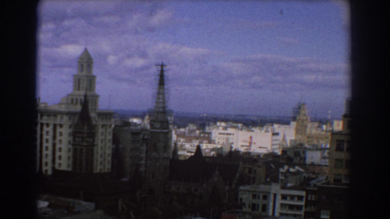 Elevated city view with church spire, towered building, rooftops, and cloudy blue sky.