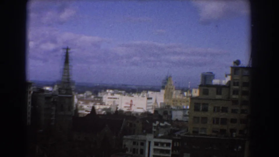 1967 Melbourne elevated view with an antenna tower, partial church spire, layered rooftops, and a taller block at right.