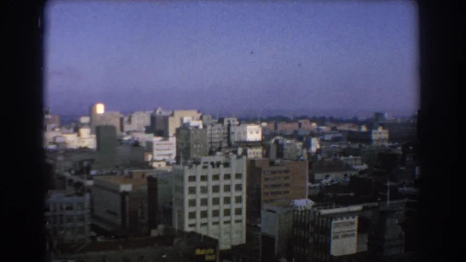 1967 Melbourne city skyline seen from above, with mid-rise buildings, rooftops, and a hazy blue sky framed by 8mm film borders.