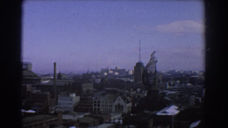Wide Melbourne skyline view with a prominent spire or tower above lower buildings, seen through soft 8mm color footage.