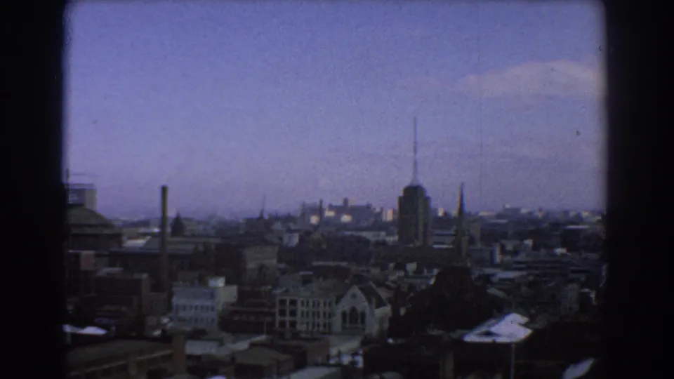 1967 Melbourne skyline from an elevated window-framed viewpoint, with rooftops, spires and a tall antenna-topped tower visible.