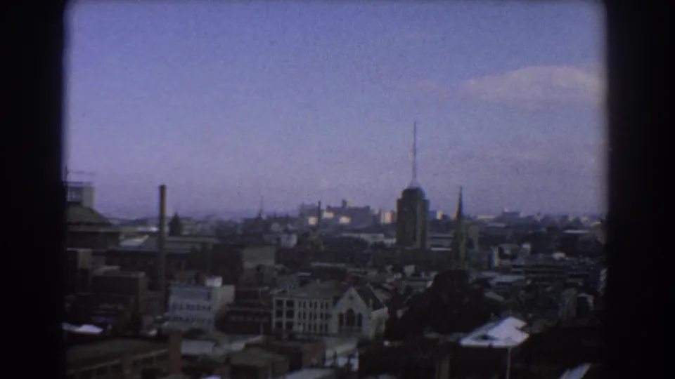 Soft, grainy 1967 Melbourne skyline view through dark framing, with rooflines, spires and an antenna-topped tower under a pale sky.