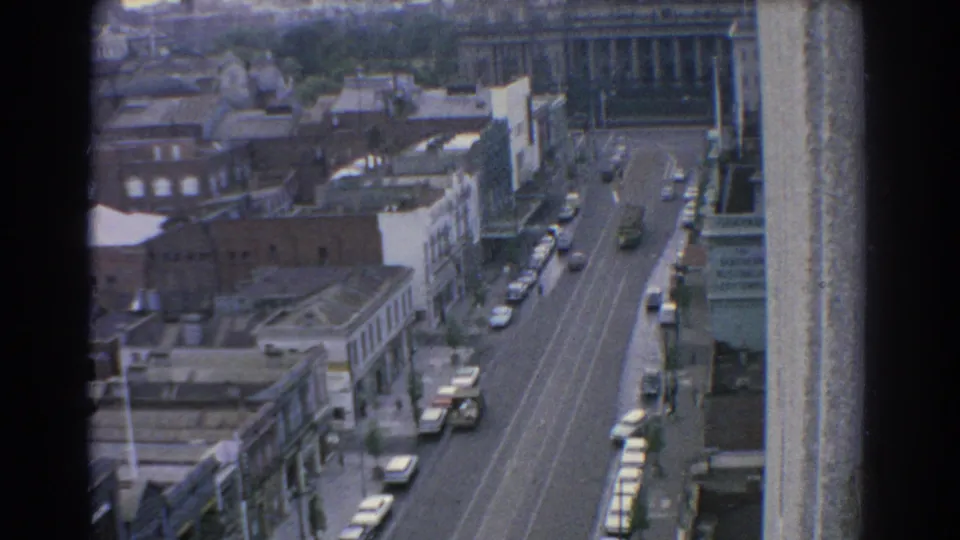 1967 Melbourne home-movie frame looking down a wide street with moving and parked cars, rooftops, and commercial buildings.
