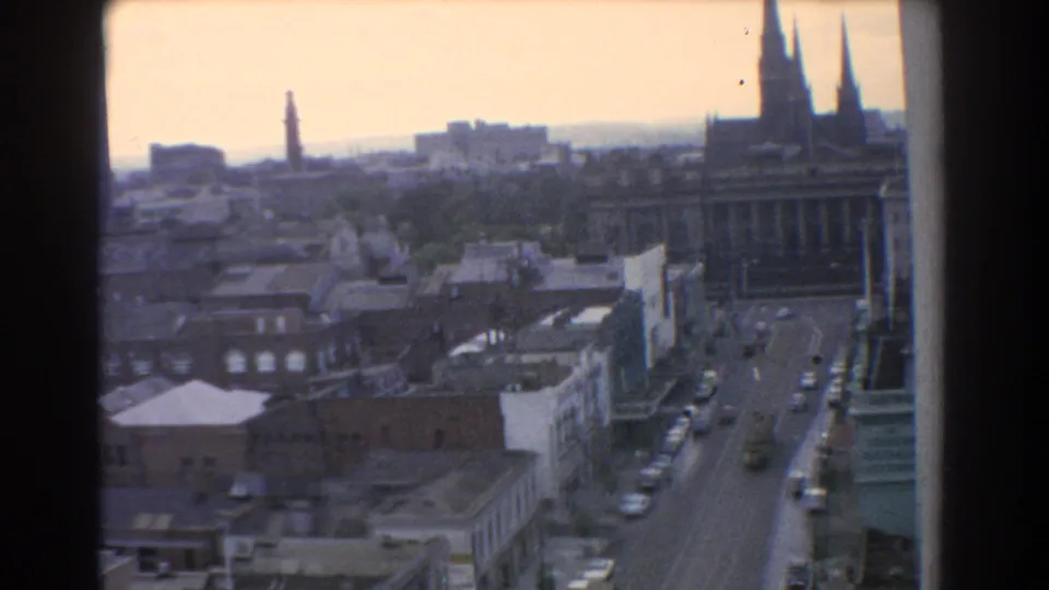 1967 Melbourne frame with a broader skyline view, street traffic below, distant tower form, and tall spires near the right edge.