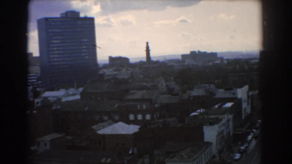 1967 Melbourne home-movie frame showing a tall office block, distant tower, low rooftops, and the edge of a busy city street.