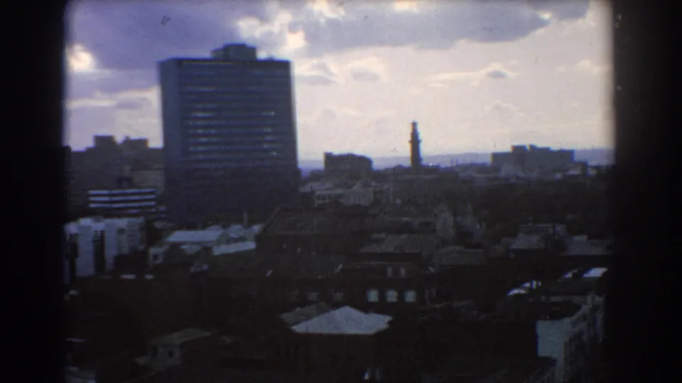 1967 Melbourne cityscape from above, with a dark high-rise block, low rooftops, distant tower, and cloudy sky.
