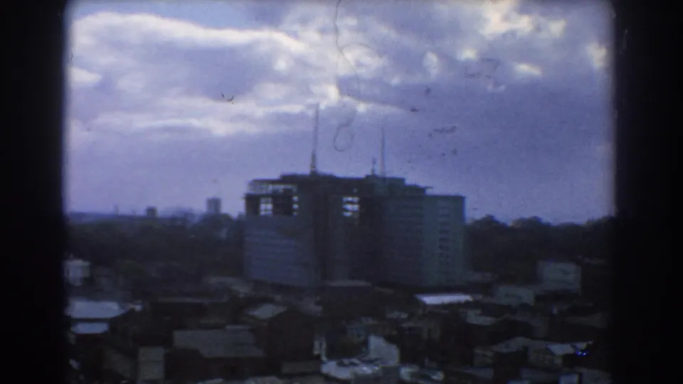 Elevated 1967 Melbourne skyline view with a prominent block-like building, rooftop antennas, shadowed rooftops, and blue-purple sky tones.