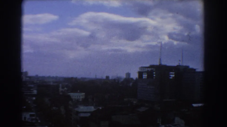 1967 Melbourne seen from above, with shadowed rooftops, a distant skyline, a tall building at right, and heavy cloud patterns across the sky.