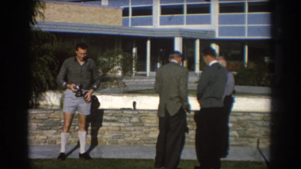 In 1967 Perth, four adult men stand outside a modern building, with one in shorts and knee-high socks holding a camera.