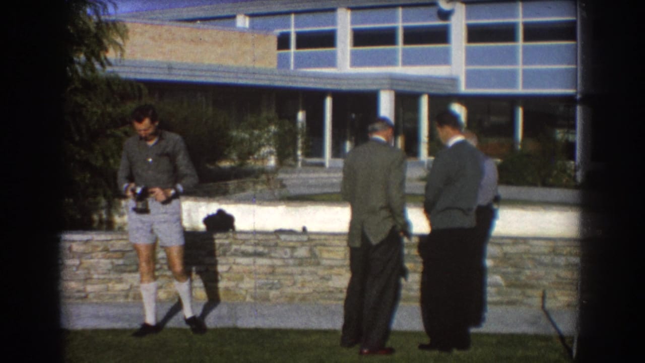 Men standing on a lawn near a stone wall and modern building, with one man holding a camera.