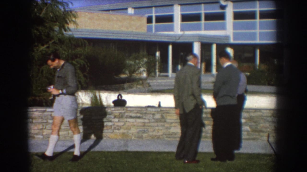 A man with a camera stands apart while two suited men face away near a stone wall.