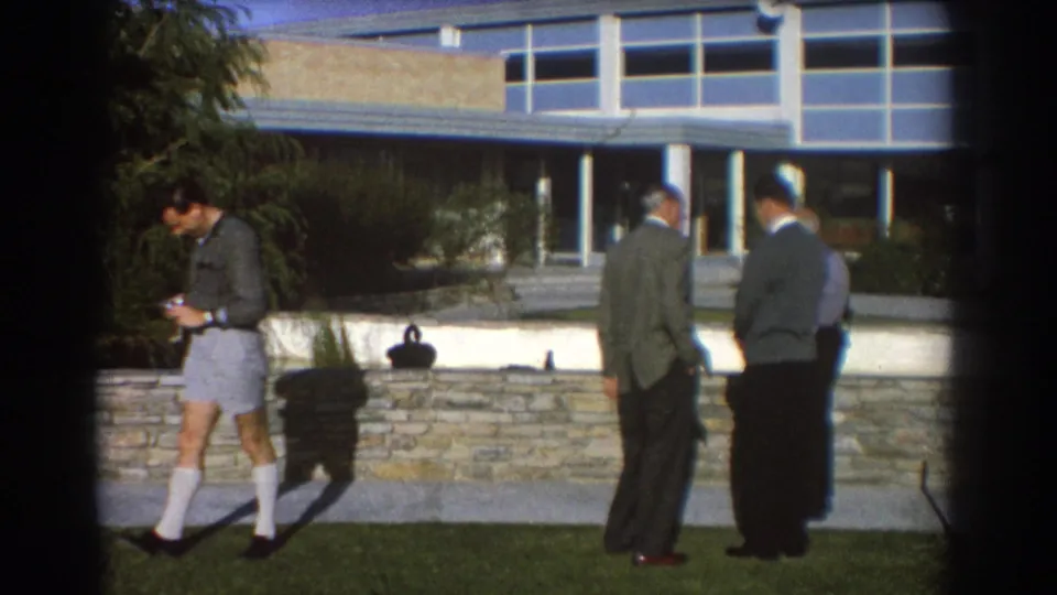 1967 Perth exterior frame showing a camera-holding man and suited men on a lawn beside a low stone wall and modern facade.