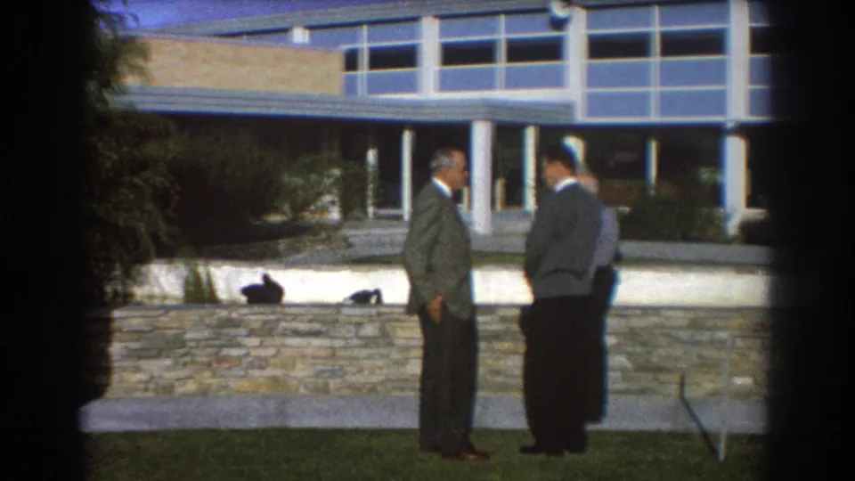 1967 Perth 8mm frame of two adult men in suits talking on a lawn outside a modern building with broad windows.