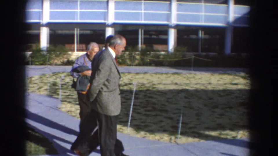 1967 Perth exterior frame of two adult men walking past a water feature, stone landscaping, and modern building windows.