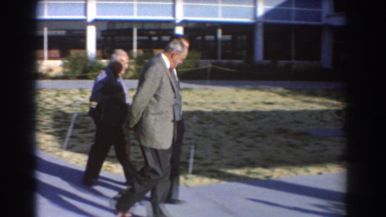 Three older men in suits walk along a paved path outside a glass-fronted building.