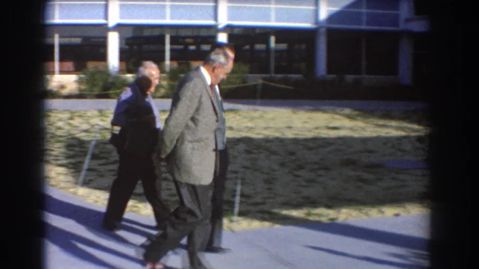 Three older white men in 1960s suits walk together outside a modern building in Perth, Australia, as seen in 8mm home-movie footage.
