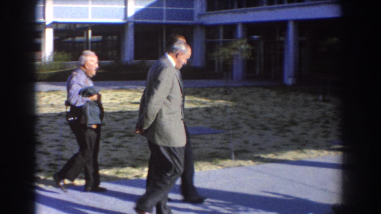 Two suited men and another man walk along a path beside a grassy area and modern building.