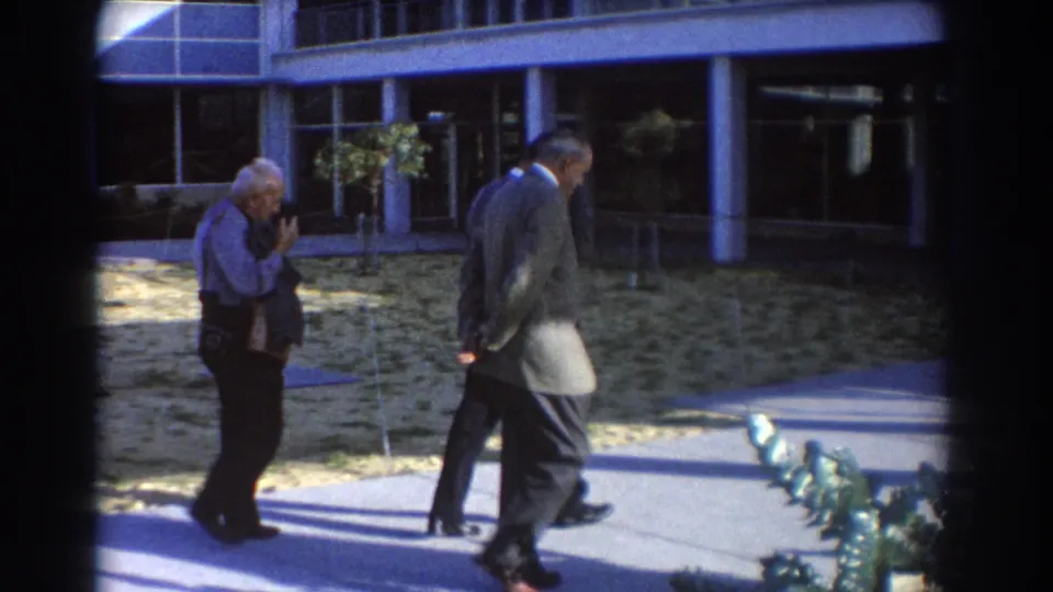 Three older men in formal clothing pass landscaping and a building entrance area in 1967 Perth home-movie footage.
