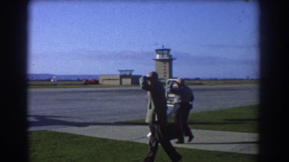 The men move near an airport apron or roadway in Perth, with distant control towers and open sky visible beyond them.