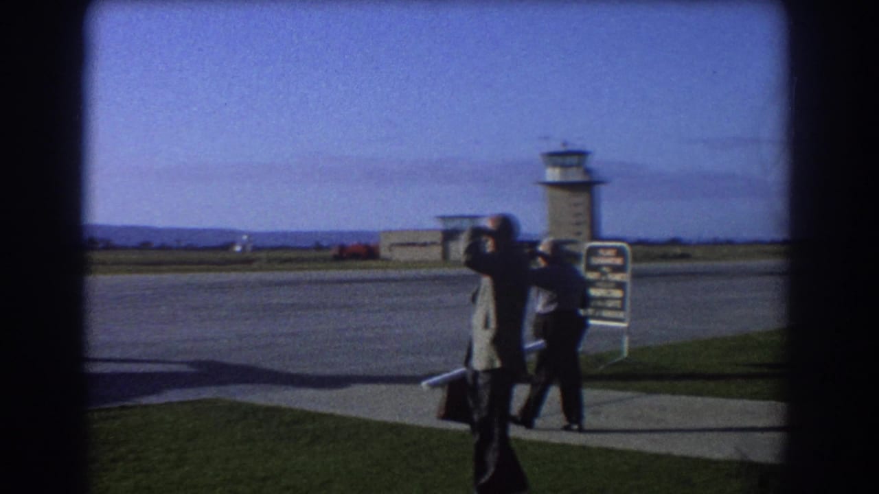 Two men near an airport tarmac with a control tower in the background.