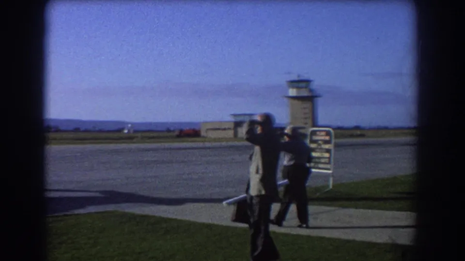 1967 Perth airport scene with two men by the runway area, one holding binoculars, with a control tower beyond the tarmac.