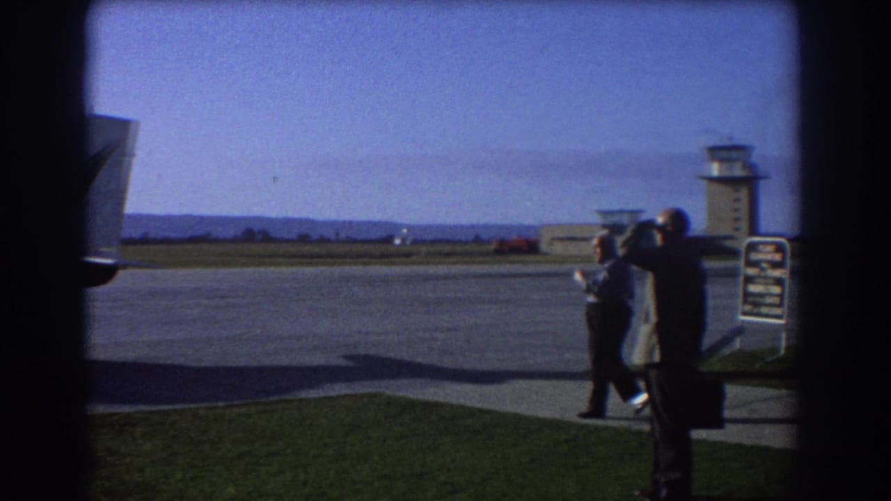 Men beside the tarmac, with one looking through binoculars and a control tower behind them.