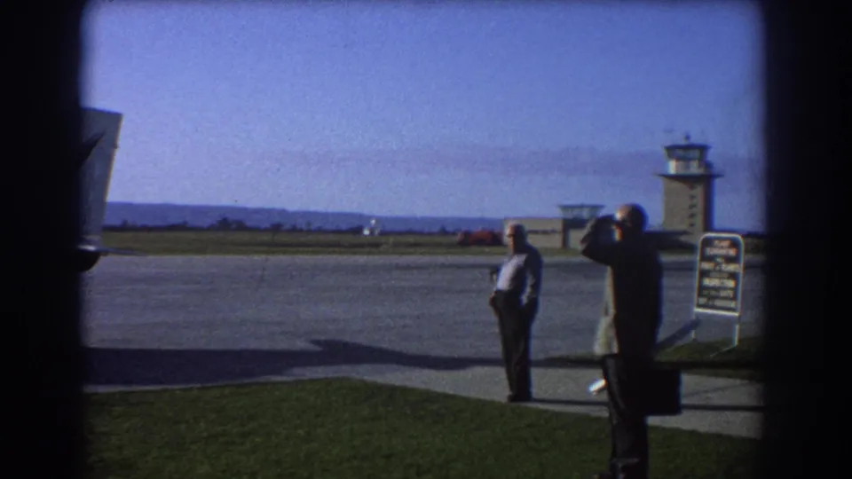 1967 home-movie frame from Perth showing a man using binoculars near the tarmac, with another man and the control tower visible.