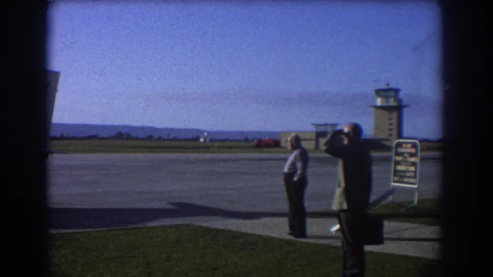 Final frame of the 1967 Perth airport view, with two men by the runway area, a sign, and the control tower across the tarmac.