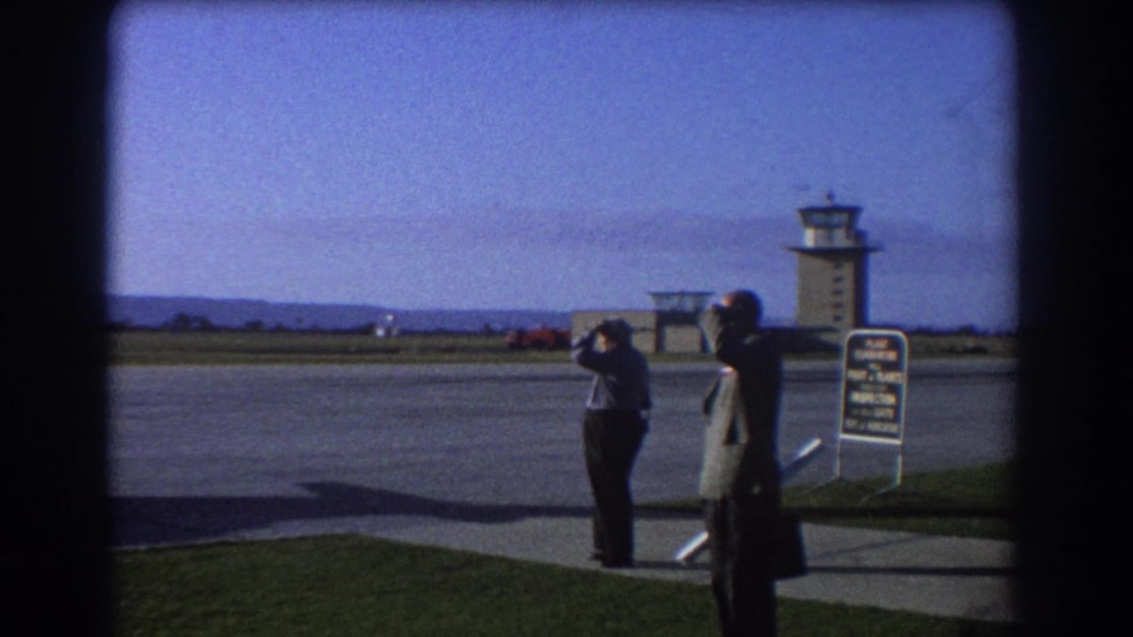 Two men stand beside an airport tarmac with a control tower and quarantine sign nearby.