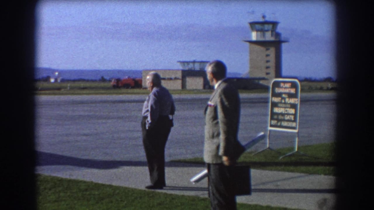 Two men stand near a runway edge with a quarantine sign and airport control tower in view.