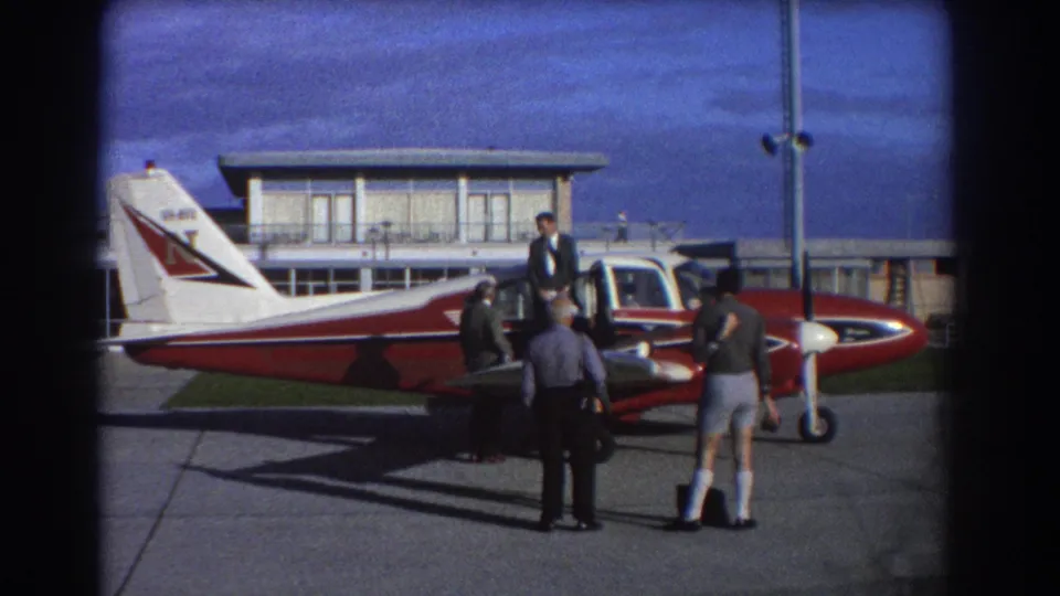 1967 Perth airfield frame with men in business and casual clothing gathered near the cockpit and nose of a red and white plane.