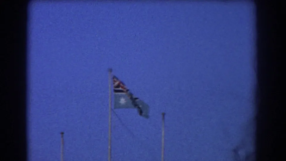 1967 Perth home-movie frame showing an Australian flag and another flag waving strongly on airfield flagpoles.