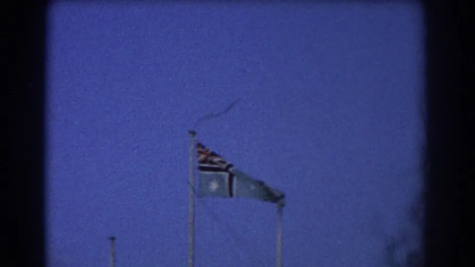 1967 Perth airfield frame focused on flagpoles, including an Australian flag, moving in visible wind under a blue sky.