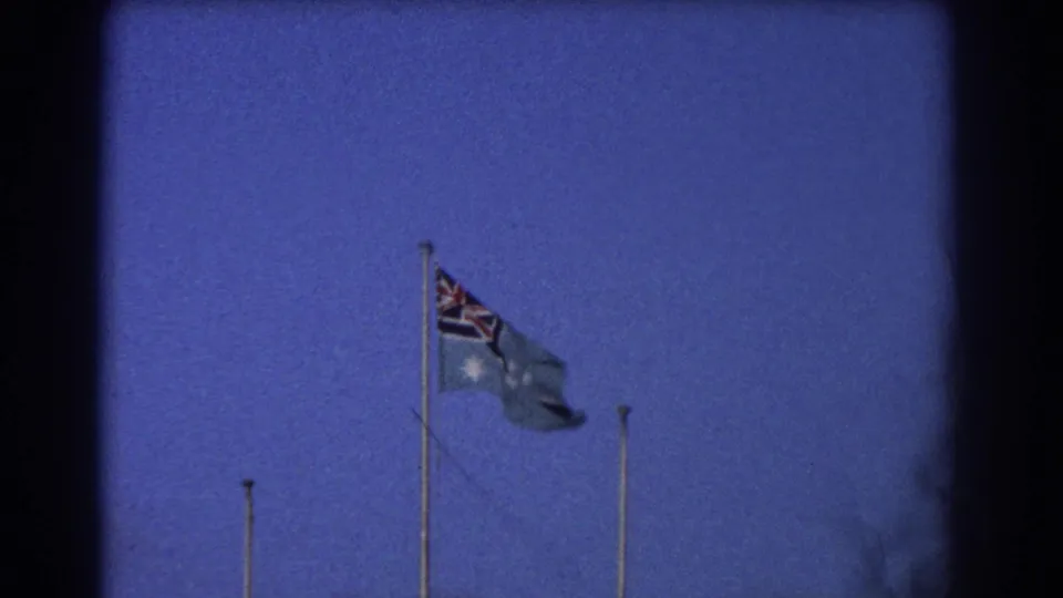 1967 Perth home-movie frame showing an Australian flag waving on a tall pole against a twilight blue sky.