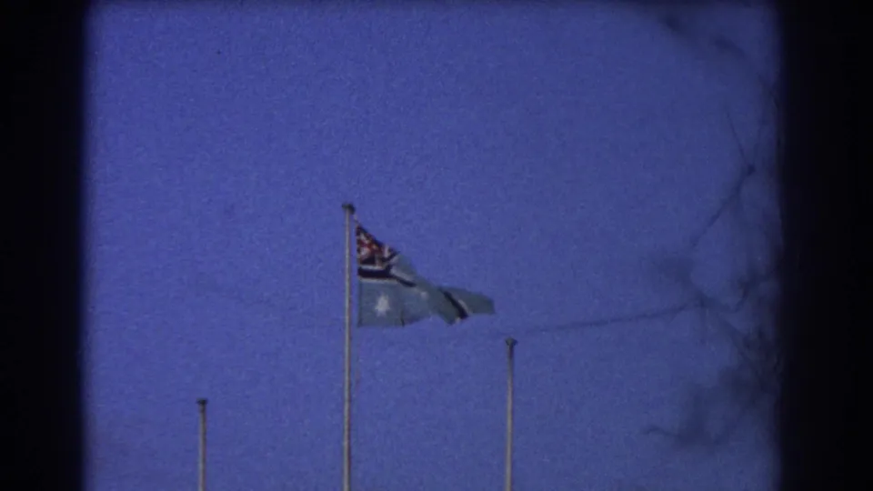 1967 Perth frame with the Australian flag extended by wind beneath a deep blue evening sky.