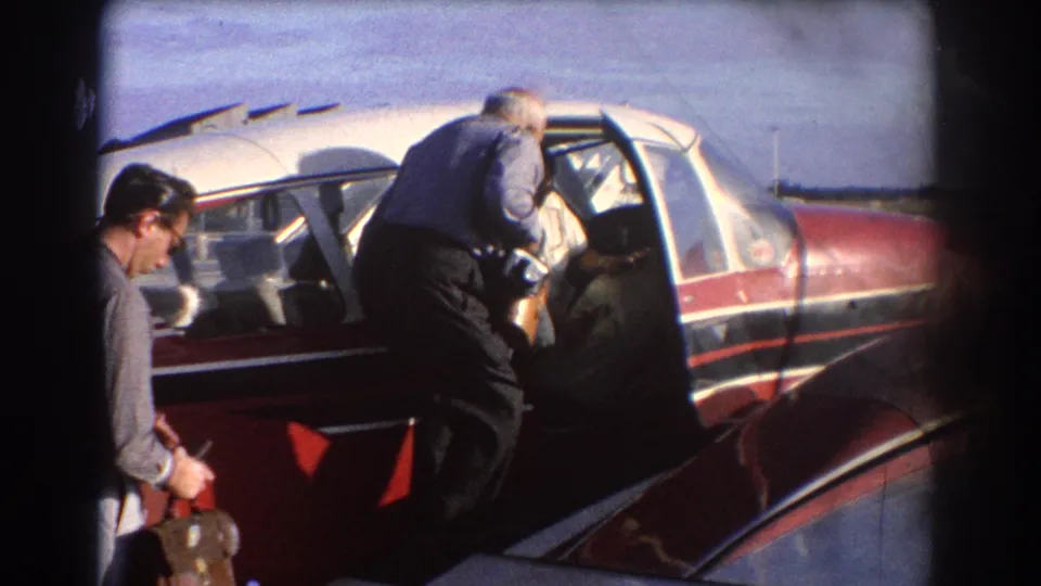 Two adult men stand beside a red and white small aircraft in Perth, with one man leaning toward the open cabin door.