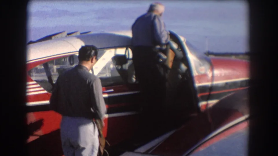 1967 Perth home-movie frame showing two passengers beside a red-and-white private aircraft at an airfield.