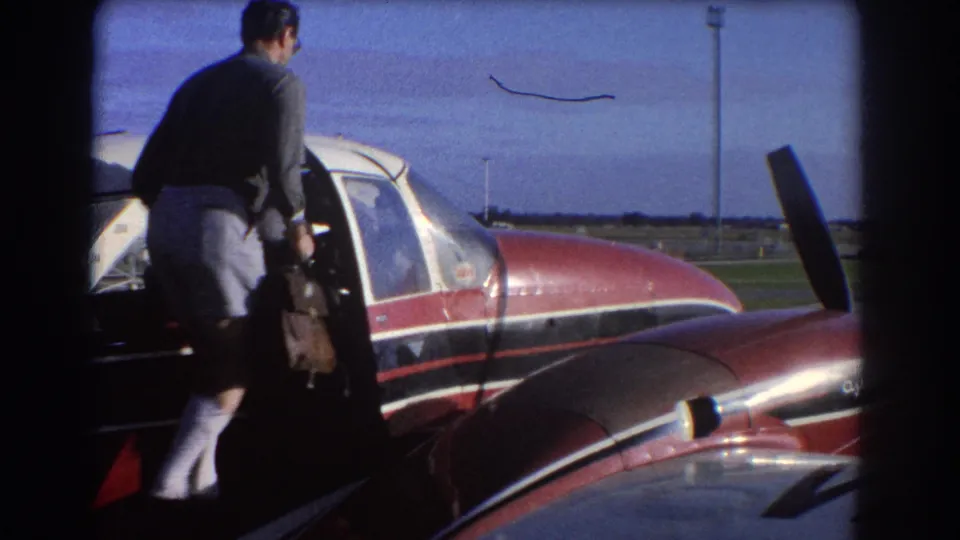 Home-movie frame from 1967 Perth showing a man loading luggage at the door of a red private aircraft.