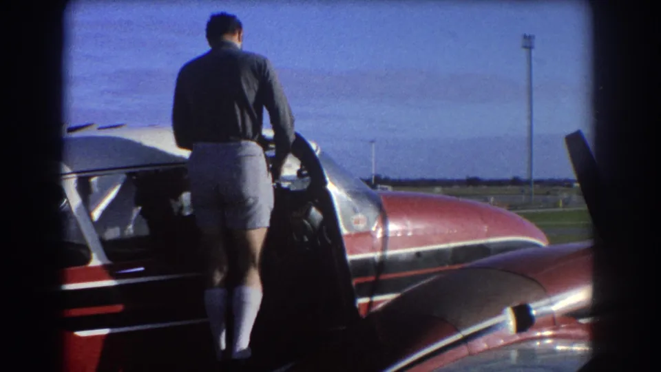 At a Perth airfield in 1967, a man stands beside the open door of a red and white small aircraft under a blue sky.
